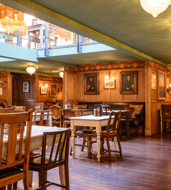 Cozy restaurant interior with wooden tables and chairs, framed portraits on wood-paneled walls, and warm ambient lighting. Sunlight streams in from a skylight above, creating a welcoming atmosphere.