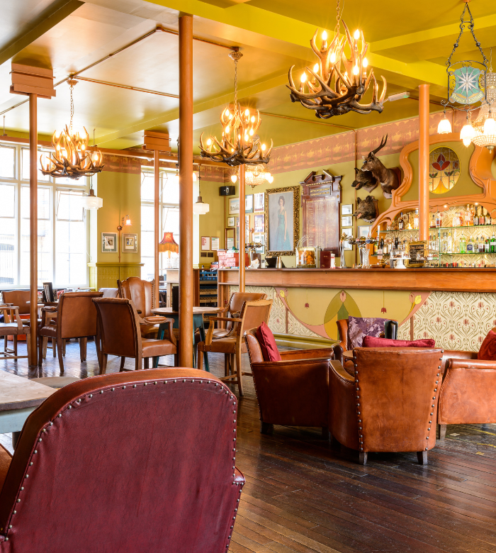 A cozy bar with leather armchairs, wooden floors, antler chandeliers, and a decorated bar counter. The warm lighting and eclectic decor create a welcoming, vintage atmosphere. Bottles and glasses line the shelves behind the bar.