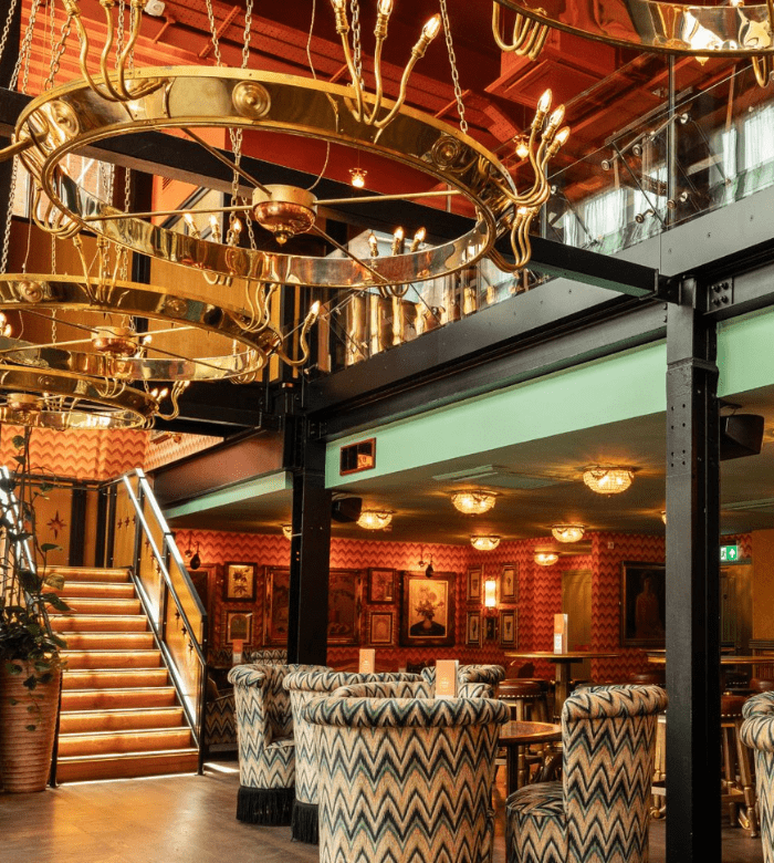 Chic restaurant interior with patterned armchairs, framed artwork on orange patterned walls, golden chandeliers, and a staircase leading to an upper level with glass panels. Warm lighting creates a cozy atmosphere.