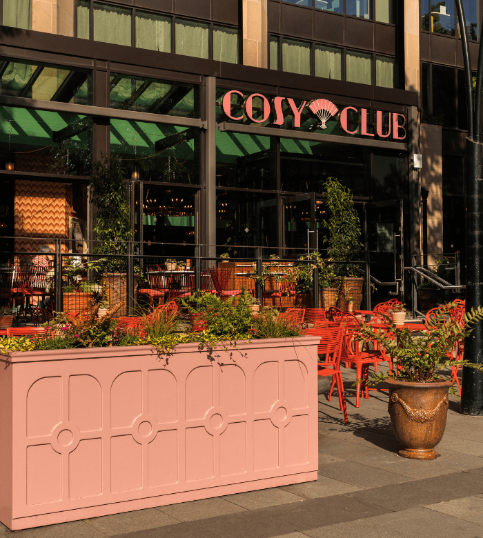 Outdoor seating area of Cosy Club restaurant with red chairs and tables, surrounded by green plants in planters, on a sunny day. Large windows and green canopy are visible in the background.