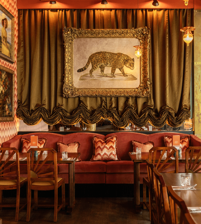 Elegant restaurant interior with a large framed painting of a leopard above a plush red velvet booth, decorative pillows, wooden chairs, and warm lighting, creating a luxurious and cozy atmosphere.