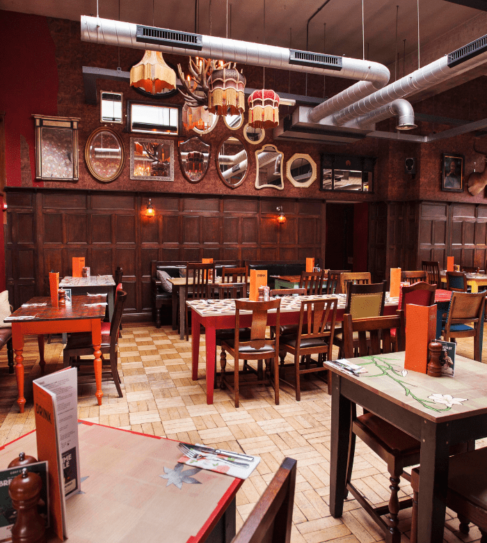 A cozy restaurant with mismatched wooden tables and chairs, brick walls, and various decorative mirrors hung above wood paneling. Menus and place settings are arranged on the tables, and warm lighting fills the space.