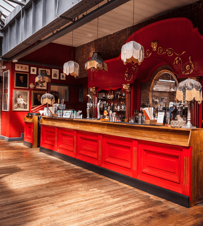 A stylish bar with bright red paneling, ornate mirrors, and vintage hanging lamps. The back wall is decorated with framed portraits, and sunlight streams in through a glass ceiling. The bar top is wooden, with bottles and glasses on display.