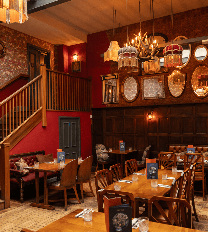 A cozy restaurant interior with wooden tables and chairs, vintage lamps, decorative mirrors on the wall, patterned wallpaper, and a staircase leading to an upper level. Place settings and menus are arranged on the tables.