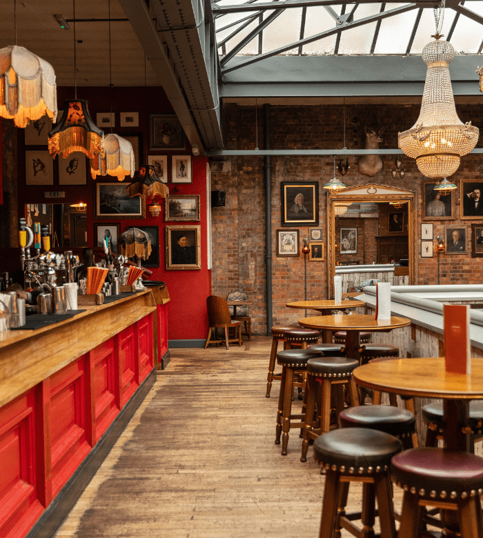 A cozy bar with red paneling, wooden floors, round tables, and leather stools. Chandeliers and vintage lamps hang from the ceiling. Framed portraits decorate the exposed brick walls. Natural light streams through the skylight.