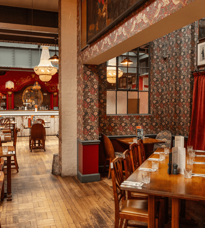 A cozy vintage-style restaurant with wooden tables and chairs, floral wallpaper, chandeliers, and a bar in the background. The space has warm lighting and eclectic decor, creating an inviting atmosphere.