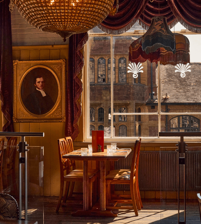 A cozy restaurant interior with a wooden table and chairs by a sunlit window, a vintage chandelier, draped curtains, a portrait painting on the wall, and a view of an old brick building outside.