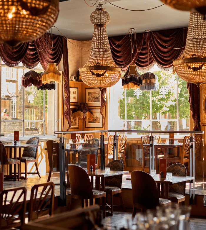 Elegant restaurant interior with large chandeliers, round tables, wooden chairs, and floor-to-ceiling windows letting in natural light, with trees visible outside. Rich, dark curtains and vintage decor create a cozy atmosphere.
