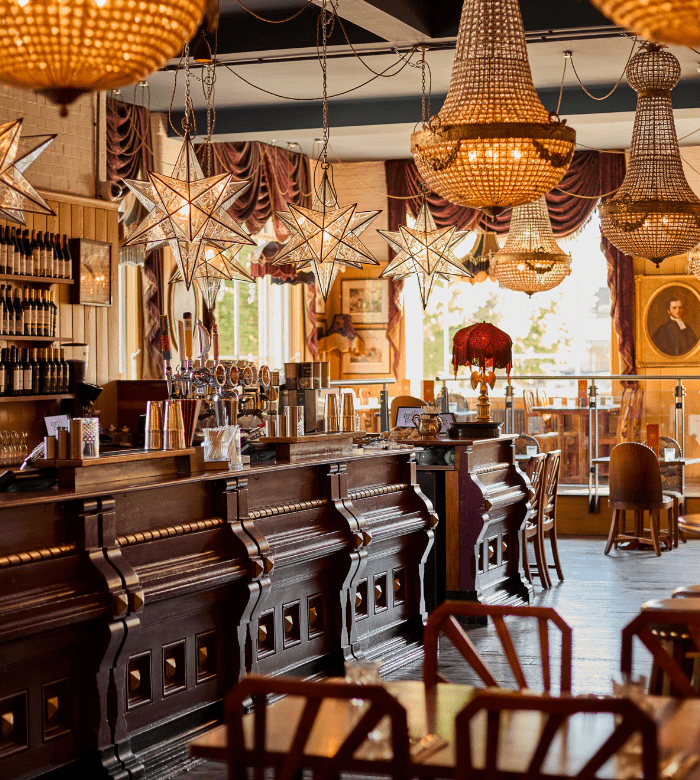 Vintage-style bar with ornate chandeliers, star-shaped hanging lights, wooden bar counter, wine bottles on shelves, velvet curtains, framed artwork, and warm sunlight streaming through the windows.