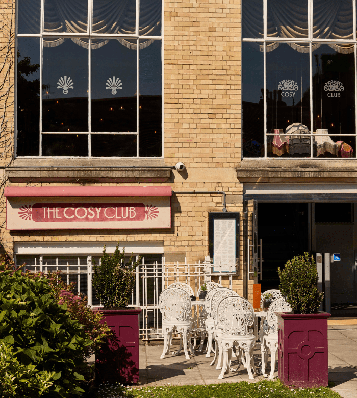 A café exterior with large windows, a sign reading “The Cosy Club,” white ornate outdoor chairs and tables, and two large planters with green shrubs in front of a beige brick building.