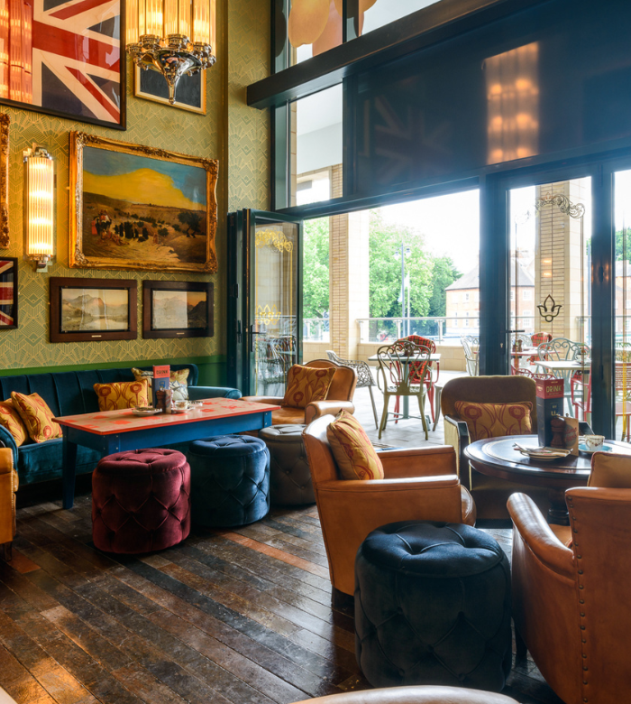 A cozy cafe with leather chairs, colorful tufted ottomans, and framed art on the walls, including a large Union Jack flag. Glass doors open to an outdoor patio with tables and chairs, overlooking greenery.