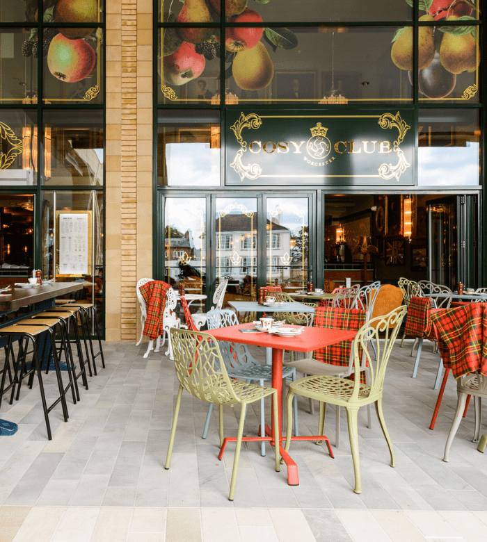 Outdoor seating area of a café with colorful metal chairs and tables, some covered with red plaid blankets, in front of large windows displaying “Cosy Club” signs and fruit illustrations. Indoor bar area is visible through the windows.