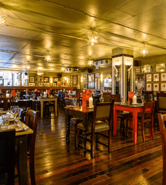 A warmly lit restaurant interior with wooden floors, tables set for dining, star-shaped pendant lights, and walls decorated with framed pictures and mirrors. The seating is a mix of chairs and booths.