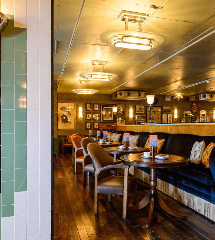 A cozy restaurant interior with wooden floors, round tables, cushioned chairs, and a long navy-blue sofa. Framed pictures decorate the wallpapered wall, and retro ceiling lights illuminate the space.