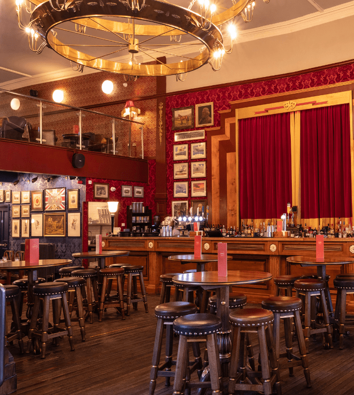 A warmly lit pub with round wooden tables and stools, a chandelier, framed photos on the wall, a long bar with taps, and a stage with red curtains in the background.