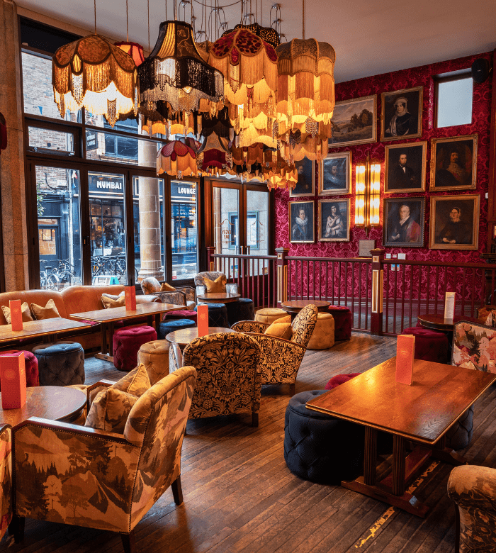 A cozy, eclectic lounge with patterned armchairs, colorful velvet couches, vintage lampshades hanging from the ceiling, and framed portraits covering deep red walls. Large windows let in natural light.