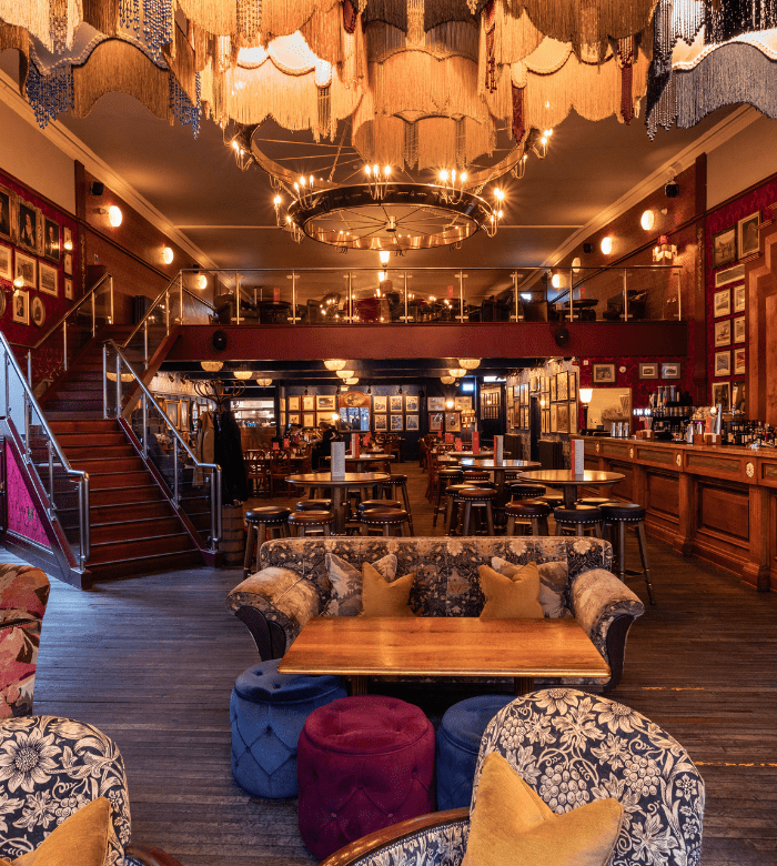 An ornate, warmly lit lounge with vintage decor, patterned sofas, plush stools, a wooden bar, and framed art on red walls. A staircase leads to an upper mezzanine level with more seating and chandeliers overhead.