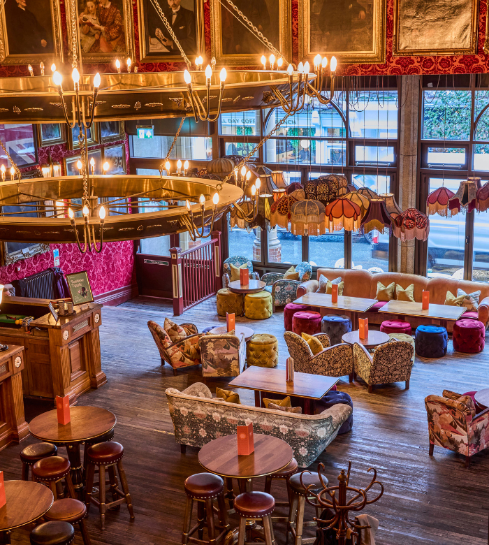 A cozy, vintage-style lounge with patterned armchairs, round tables, colorful cushions, eclectic lampshades, and large windows. Chandeliers hang from the ceiling, and paintings decorate the red walls above a polished wooden floor.