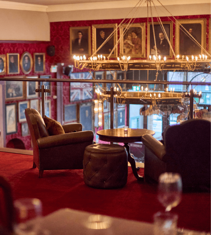 Two leather armchairs and a small table sit on a red carpeted balcony in an elegant lounge, with framed portraits on the walls, a glass railing, and a large chandelier overhead.