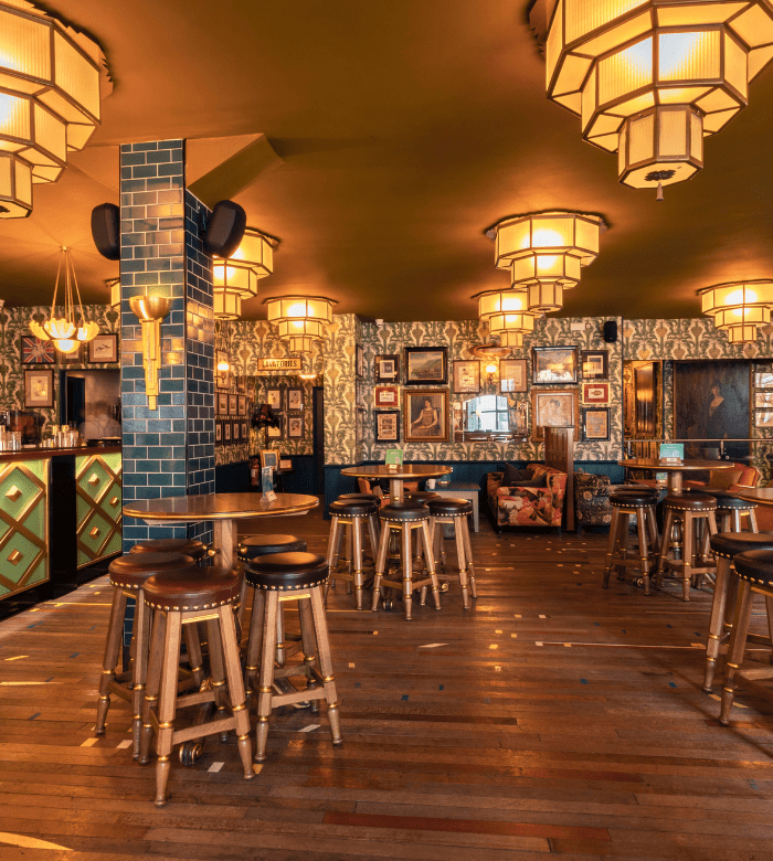 A stylish bar interior with patterned wallpaper, framed art, round wooden tables with stools, green tiled columns, and warm, geometric ceiling lights creating a cozy, retro atmosphere.