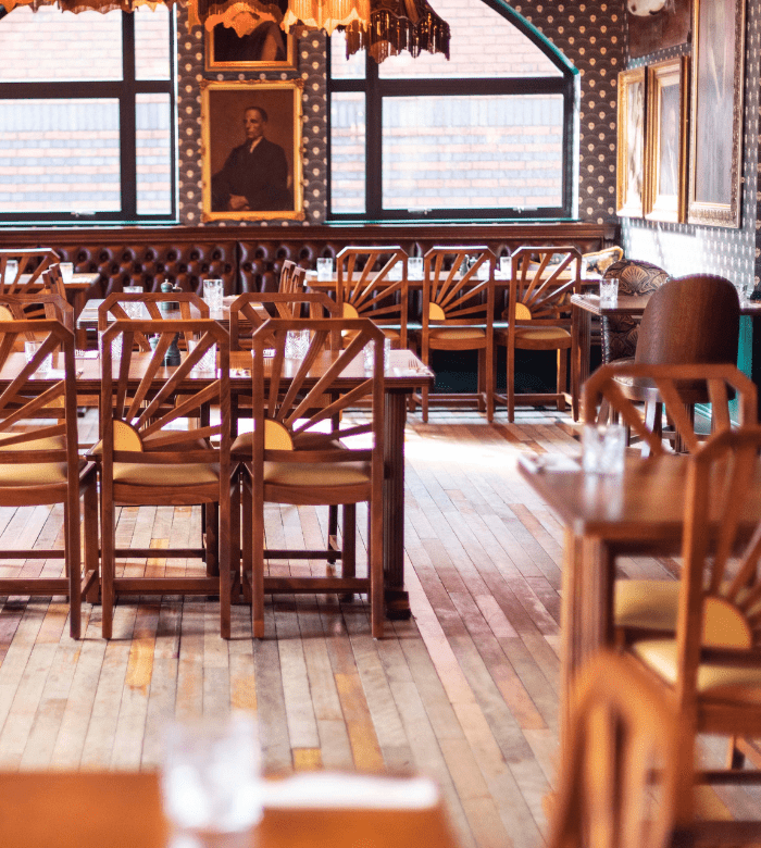 A cozy, empty restaurant with wooden chairs and tables, glasses set on each table, patterned wallpaper, large windows, and framed portraits on the walls. Warm lighting creates a welcoming atmosphere.
