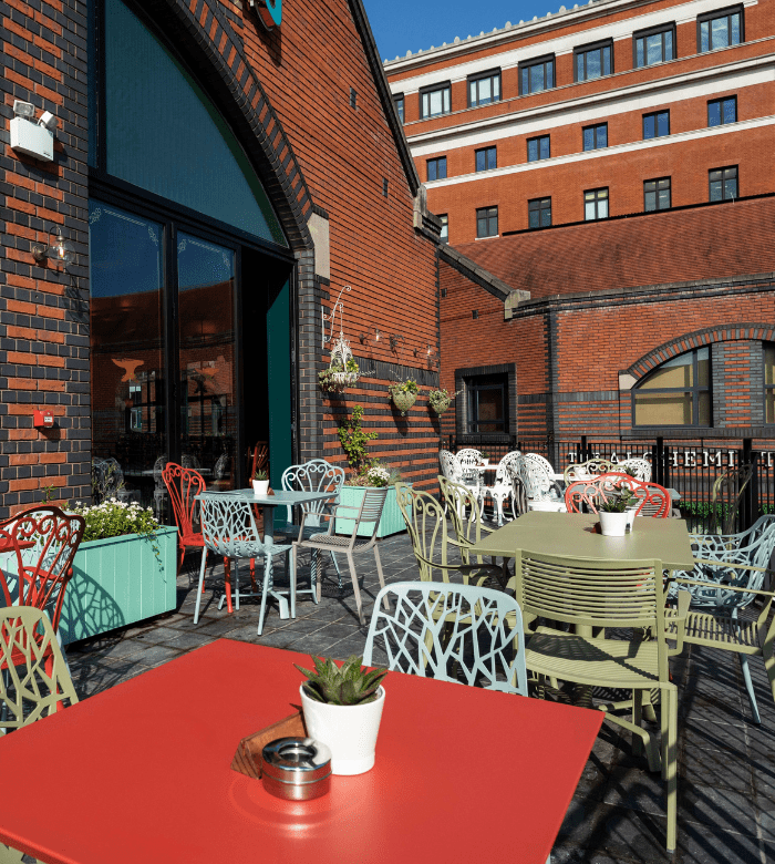 Outdoor patio with colorful tables and chairs, potted plants, and brick buildings in the background under a clear blue sky. The setting appears to be a café or restaurant terrace.