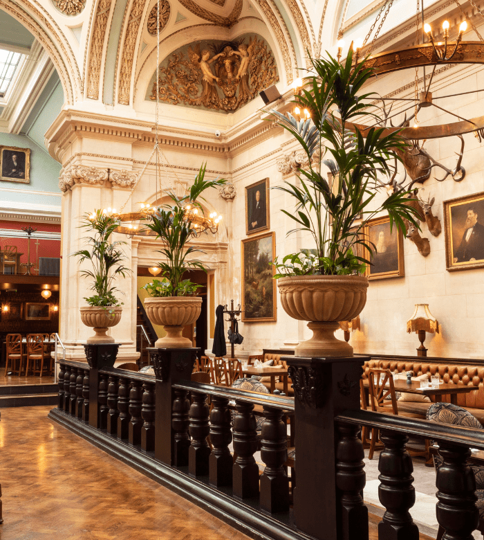 Elegant restaurant interior with high ornate ceilings, large potted plants, classic paintings on cream-colored walls, wooden floors, and leather seating, creating a sophisticated and historic atmosphere.