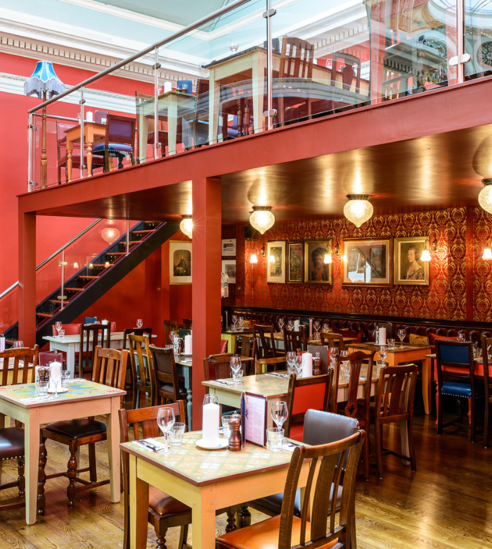 A vibrant restaurant interior with eclectic wooden tables and chairs, red walls adorned with framed art, chandeliers, and an upper mezzanine seating area visible above an open staircase.