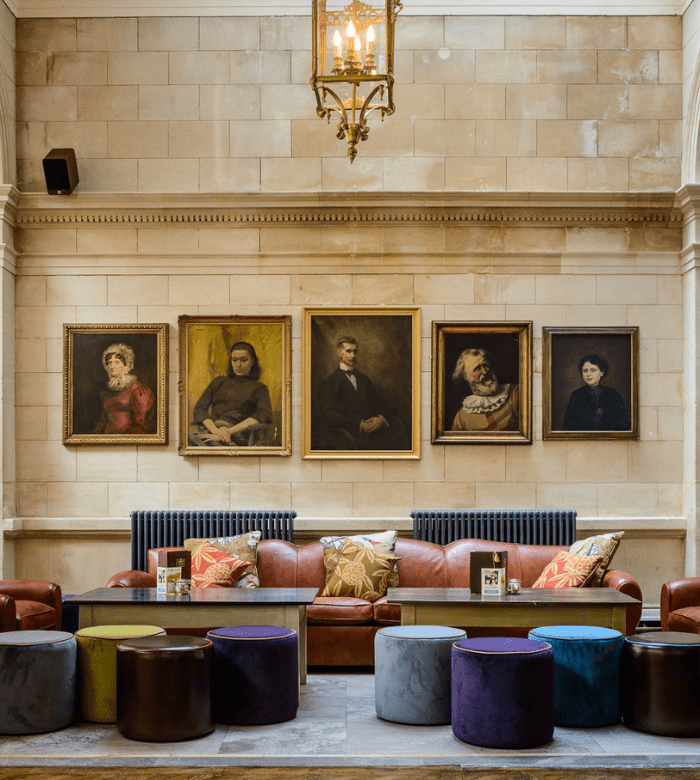 A stylish lounge with leather sofas, colorful cushions, velvet ottomans, and wooden tables. Five framed portraits hang on a stone wall above, beneath a large hanging chandelier.