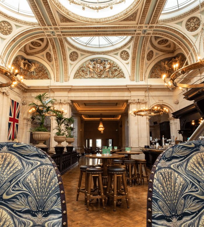 An ornate interior of a grand hall featuring a large glass dome, elaborate ceiling murals, chandeliers, a British flag, round tables with stools, and plush armchairs with blue patterned upholstery.