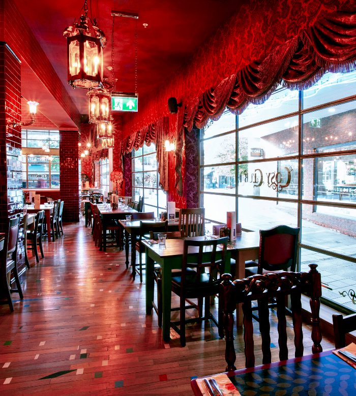 A restaurant with ornate red decor, patterned curtains, hanging lanterns, and wooden tables and chairs. Large windows line one side, letting in natural light and offering a view of the street outside.
