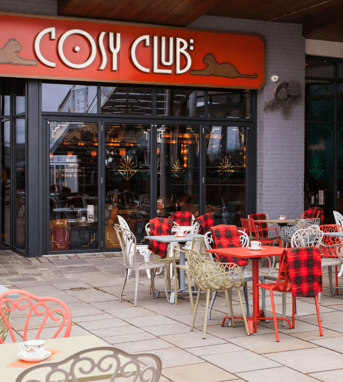 Outdoor seating area of Cosy Club café with decorative metal chairs, red tables, and tartan blankets draped over some seats. Glass doors and a red sign with "Cosy Club" and lounging cat logos above the entrance.