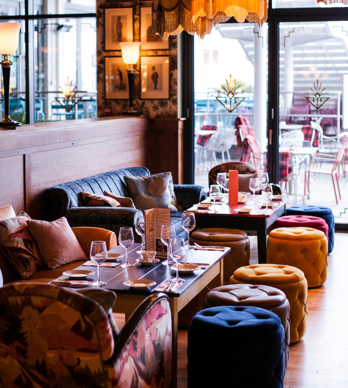 A cozy, stylish restaurant interior with plush seating, colorful ottomans, set dining tables with glasses and plates, warm lighting, and large windows leading to an outdoor patio with tables and chairs.