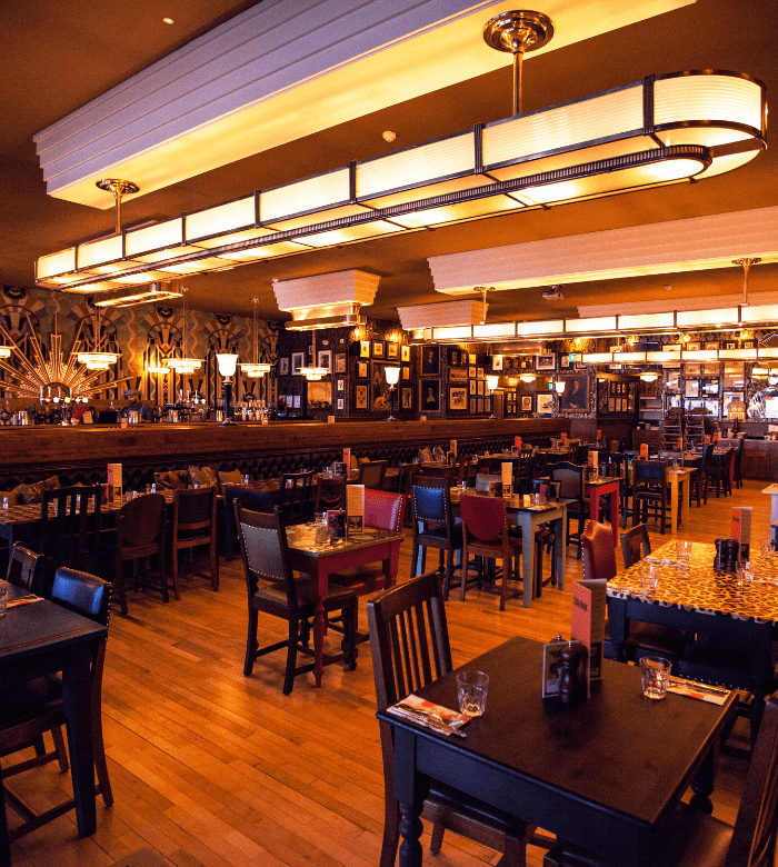 A warmly lit restaurant interior with wooden floors, dark tables and chairs, and walls decorated with framed pictures and mirrors. The tables are set for dining but the restaurant is empty of patrons.