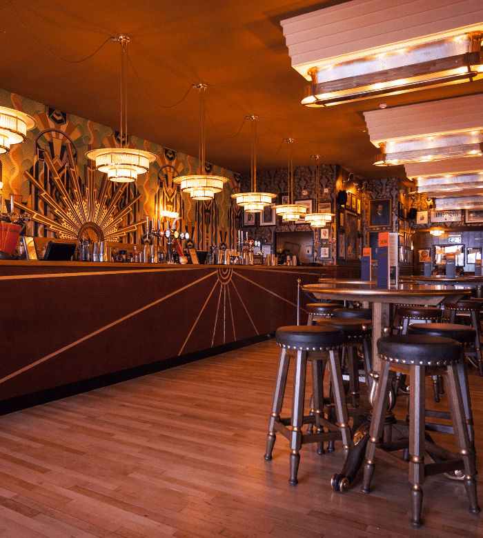 A stylish bar with art deco decor, geometric patterns on the bar front, vintage pendant lights, framed photos on the walls, high tables, and wooden stools on a polished wooden floor.