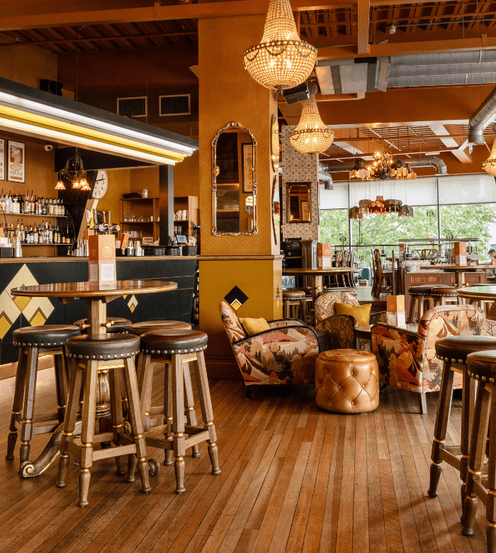 A cozy bar with wooden floors, high stools, and round tables. Vintage chandeliers hang from the ceiling. There are armchairs with floral upholstery and a gold ottoman in a relaxed seating area near large windows.