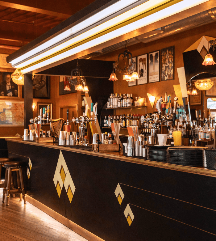 A stylish bar with geometric decor features hanging lights, various bottles, glassware, cocktail shakers, and framed art on the walls, creating a warm, inviting atmosphere.