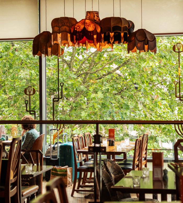 A cozy restaurant with wooden tables and chairs, retro hanging lamps, and large windows showing leafy green trees outside. A person sits alone at a table, and a suitcase is visible nearby.