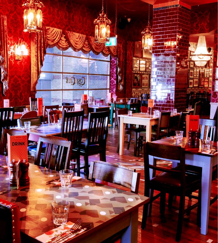 A warmly lit restaurant with patterned wallpaper, vintage decor, wooden tables and chairs, glassware on tables, and red accents creating a cozy, inviting atmosphere.