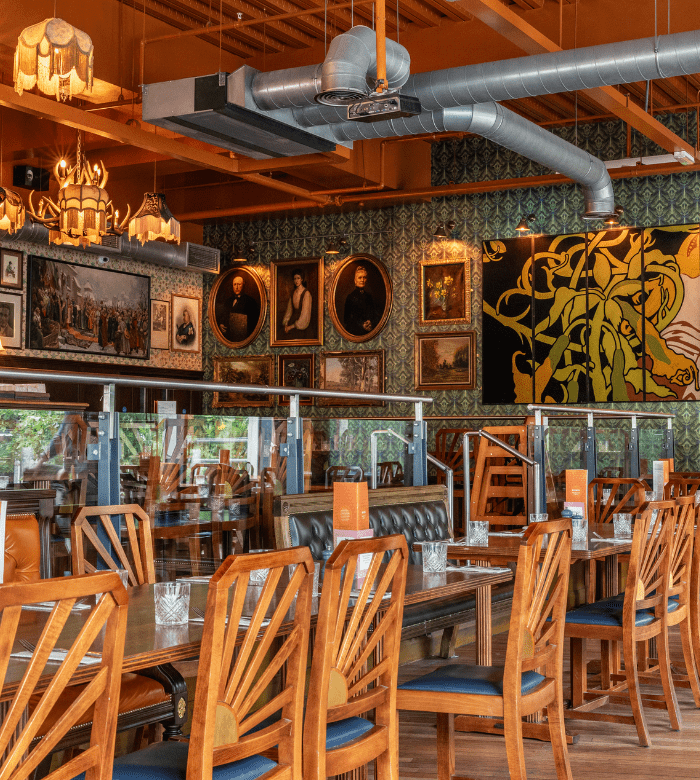 A cozy restaurant interior with wooden chairs and tables, glass dividers, eclectic wall art, framed portraits, and decorative lighting fixtures hanging from the ceiling.