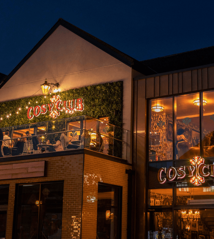 A warmly lit restaurant called "Cosy Club" at night, with outdoor seating on a balcony, string lights, and people dining inside and outside. The building exterior is decorated with greenery and modern brickwork.