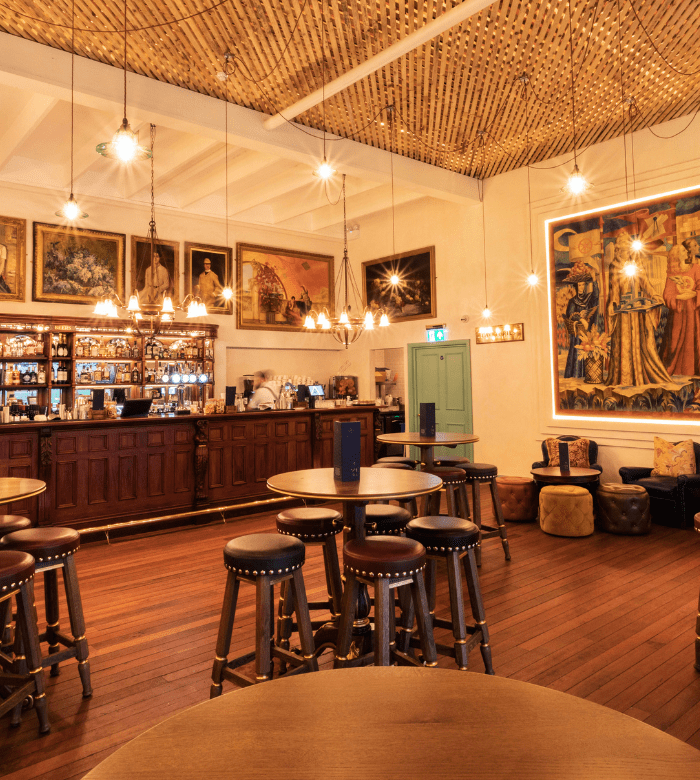 A cozy, warmly lit bar with round wooden tables, high stools, a wooden floor, and a classic bar counter. The walls are decorated with large paintings and tapestries, creating an inviting, vintage atmosphere.