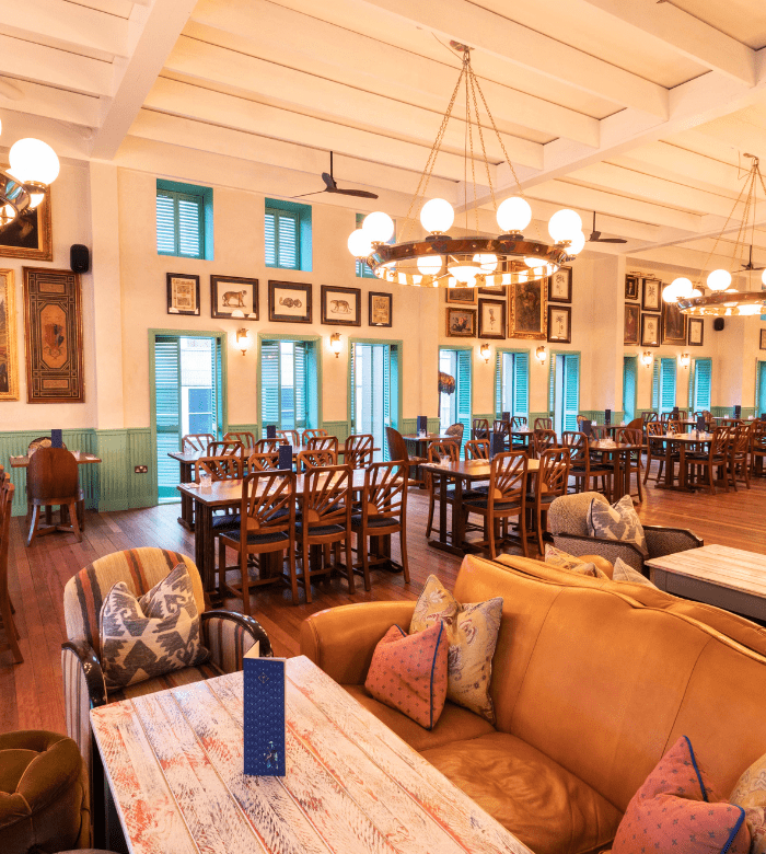 A spacious, well-lit dining room features wooden tables and chairs, soft sofas with cushions, chandeliers, framed art on cream walls, and large blue-trim windows letting in natural light.