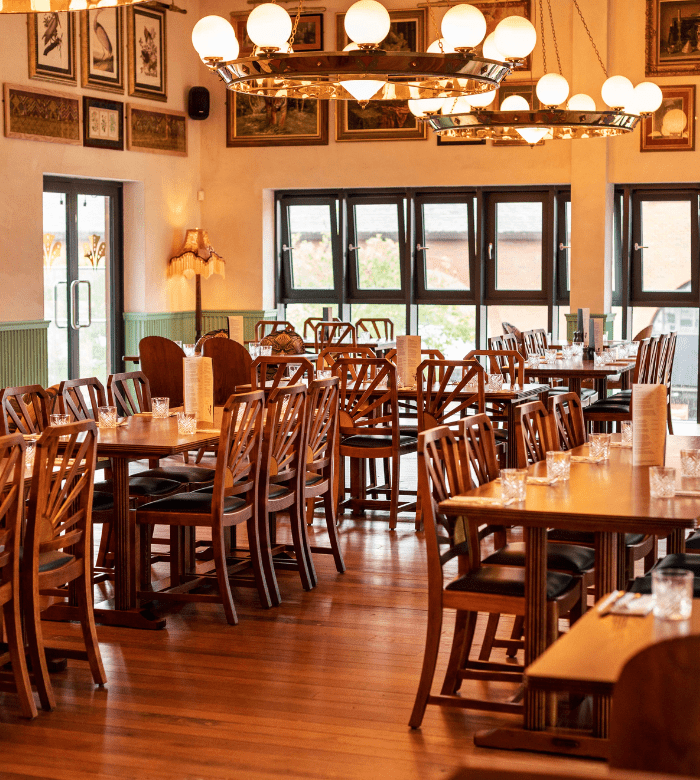 A cozy restaurant dining area with wooden tables and chairs, set for guests. Warm lighting hangs from the ceiling, and large windows let in natural light. Framed pictures decorate the cream-colored walls.
