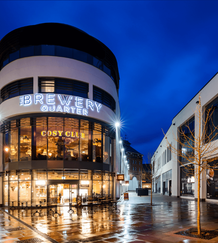 A modern, curved building at night with "The Brewery Quarter" and "Cosy Club" signs lit up; wet pavement reflects lights and leafless trees stand nearby under a deep blue sky.