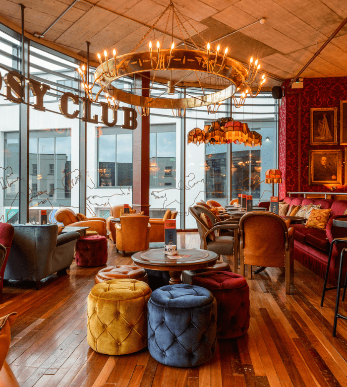 A cozy, colorful lounge with plush velvet chairs and ottomans, wooden floors, eclectic decor, chandeliers, and large windows letting in natural light. "CLUB" is visible on a window.