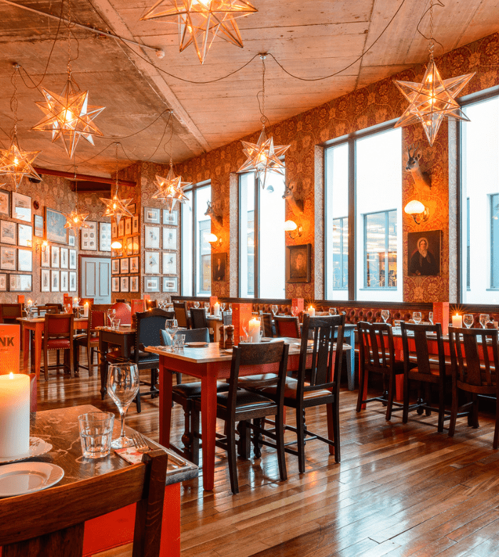 A cozy restaurant interior with wooden floors, star-shaped pendant lights, and walls decorated with framed pictures and portraits. Tables are set with glassware and candles, creating a warm, inviting atmosphere.