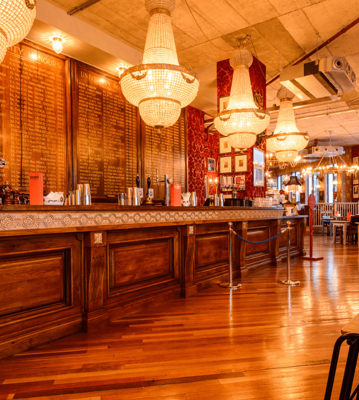 An elegant bar with large chandeliers, polished wooden floors, and a long wooden counter. The back wall features a gold-colored board with names, and red wallpaper creates a warm, inviting atmosphere.