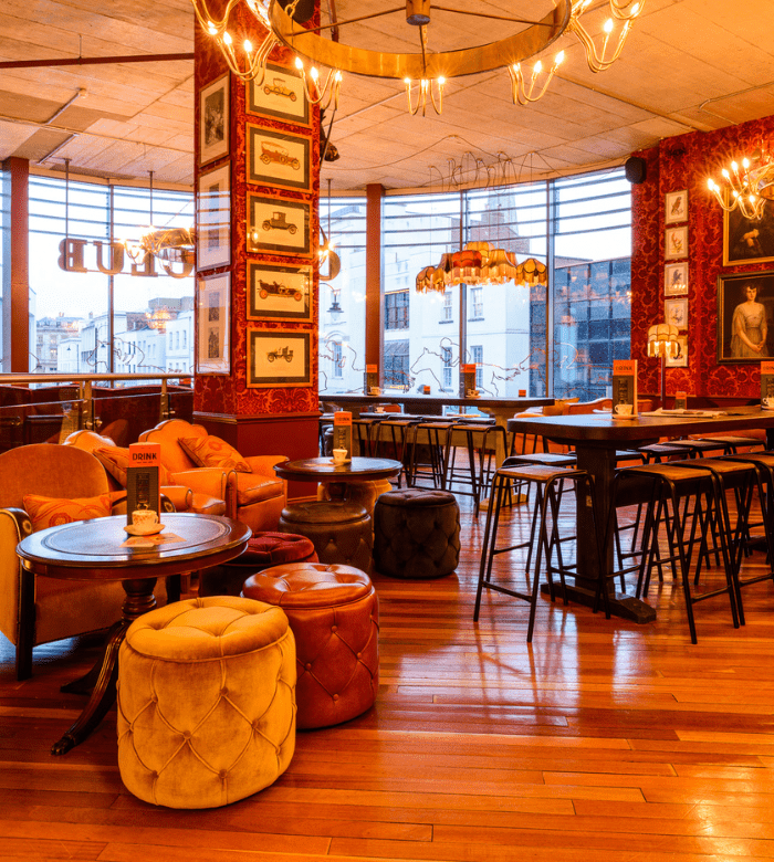A cozy, stylish restaurant interior with warm lighting, leather chairs, plush ottomans, wooden floors, high tables, and framed art on red wallpapered walls. Large windows let in natural light.