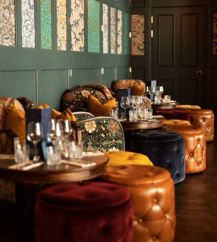 Cozy restaurant seating with plush velvet and leather chairs, round wooden tables set with glassware, and decorative wall panels with floral patterns against dark green walls. Warm lighting creates an inviting atmosphere.